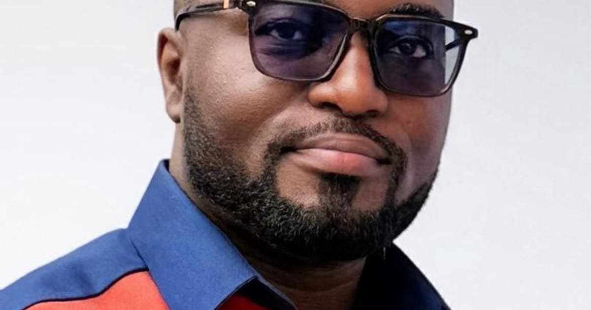 Close-up portrait of a man wearing dark sunglasses and a blue shirt with red accents, looking at the camera.