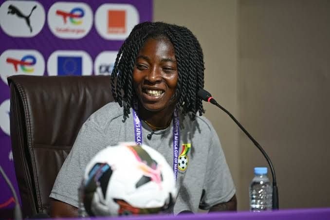 Smiling woman with braided hair sits at a press conference table, soccer ball in foreground, purple backdrop with logos behind her.