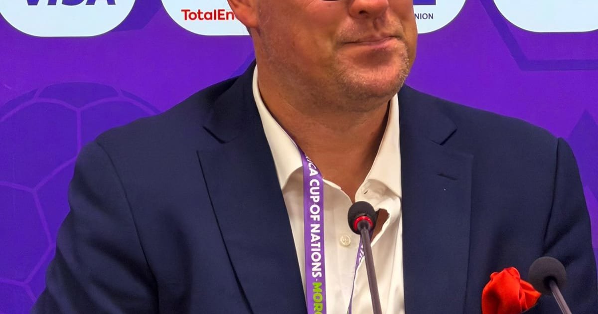 Man in a navy suit at a podium with microphones, purple event backdrop and lanyard visible